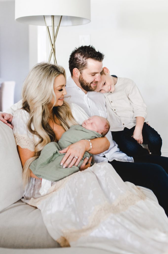 sweet family cuddle on the couch during their in-home newborn session. Big brother cuddles on dad while mom holds the newborn baby in a light green swaddle