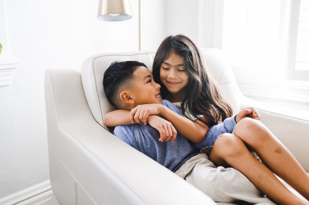 brother and sister cuddling in a white chair while light streams into the white bedroom during a family photo session at home