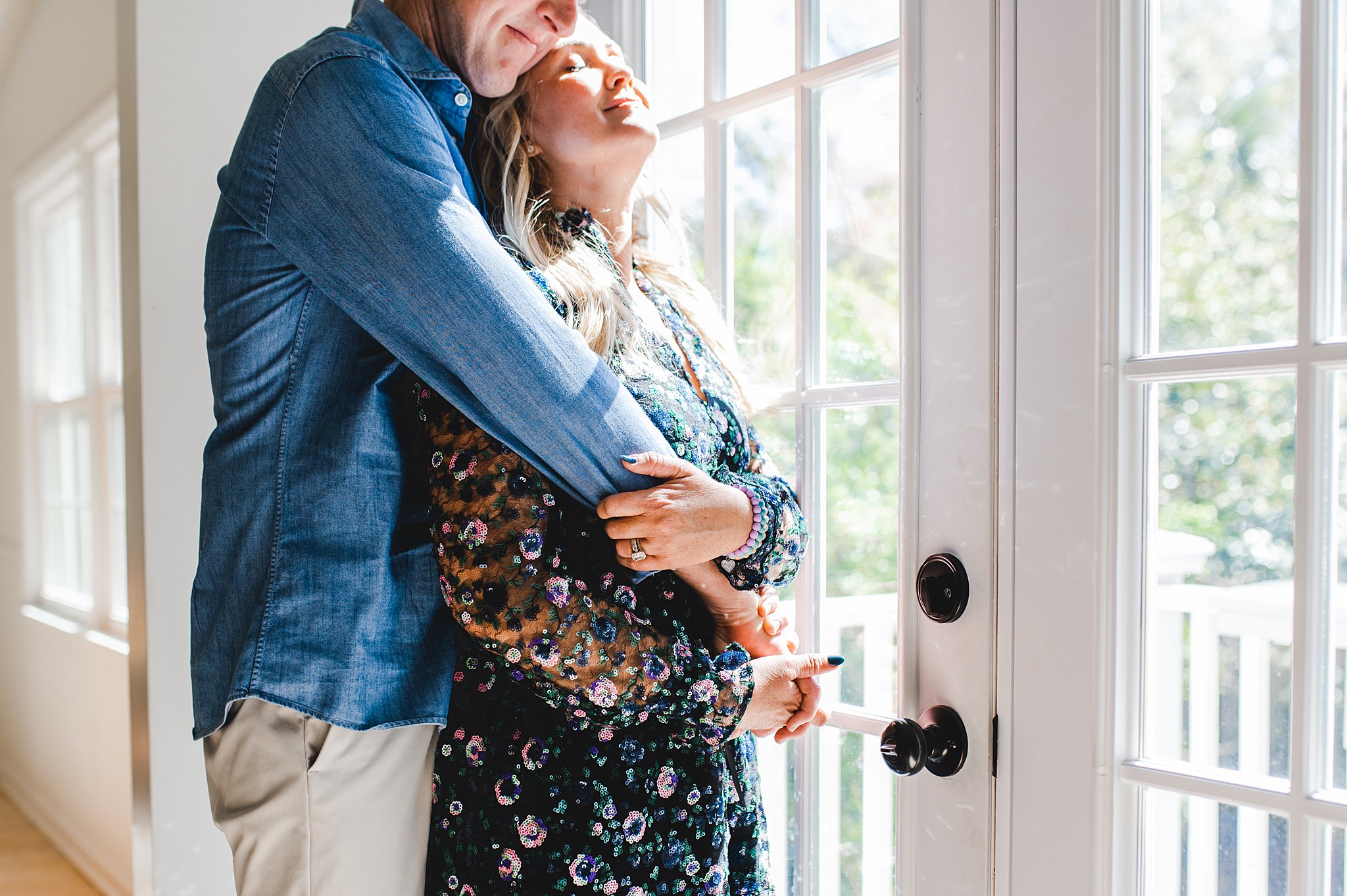 Mom wears a sequin dress for her family photos. Dad hugs her from behind near a window.