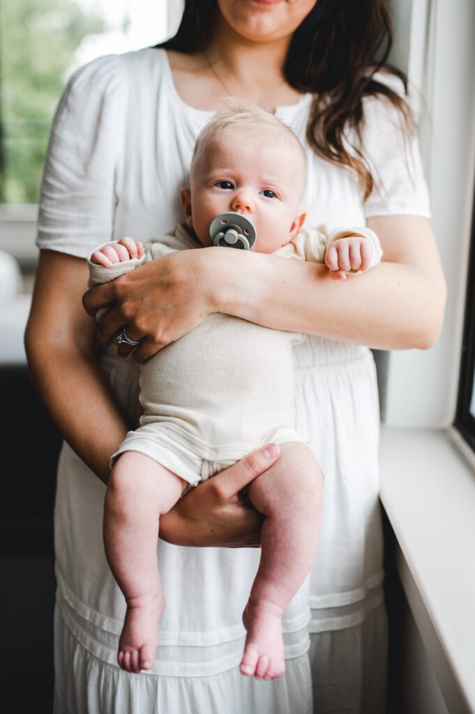 mom holding newborn baby with a pacifier next to a window with sunlight streaming in. The baby is facing the camera. This post answers FAQs about your photography session.