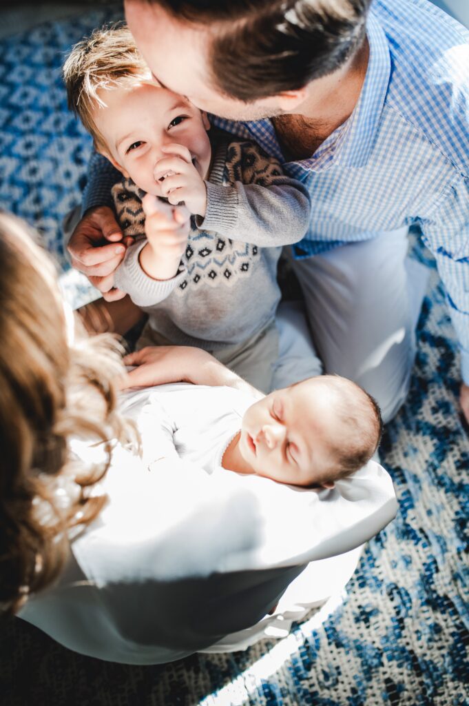 Dad kissing new big brother while mom holds newborn baby brother in a sunsoaked natural light room